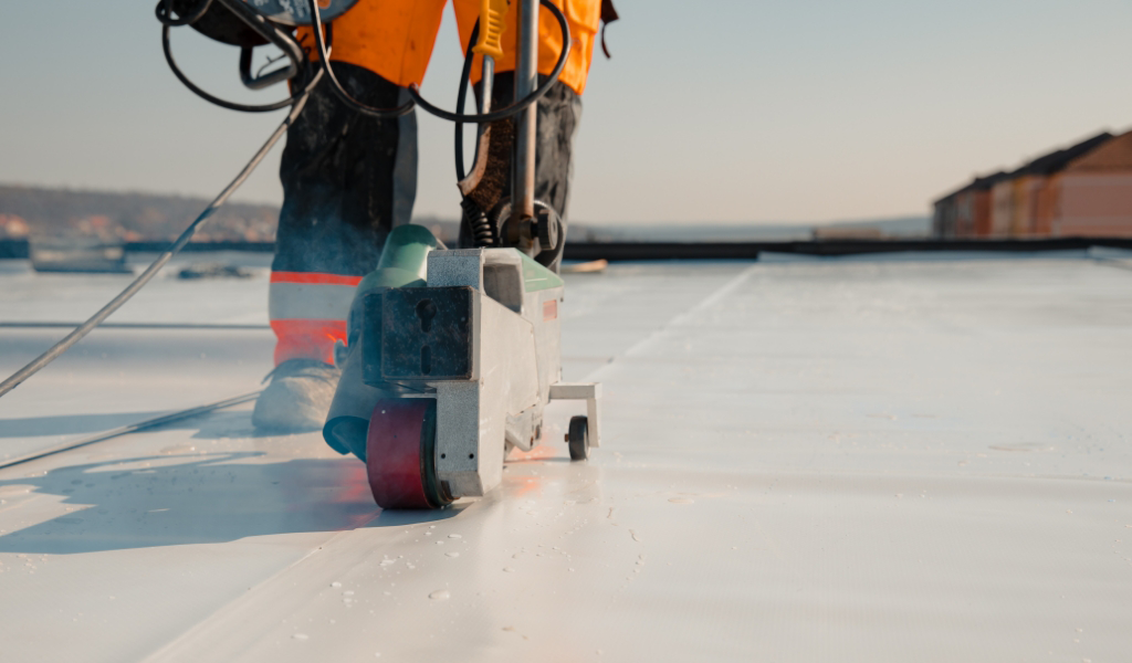 A worker in orange overalls expertly uses a machine to weld seams on a flat white roof, showcasing top-notch roofing services.