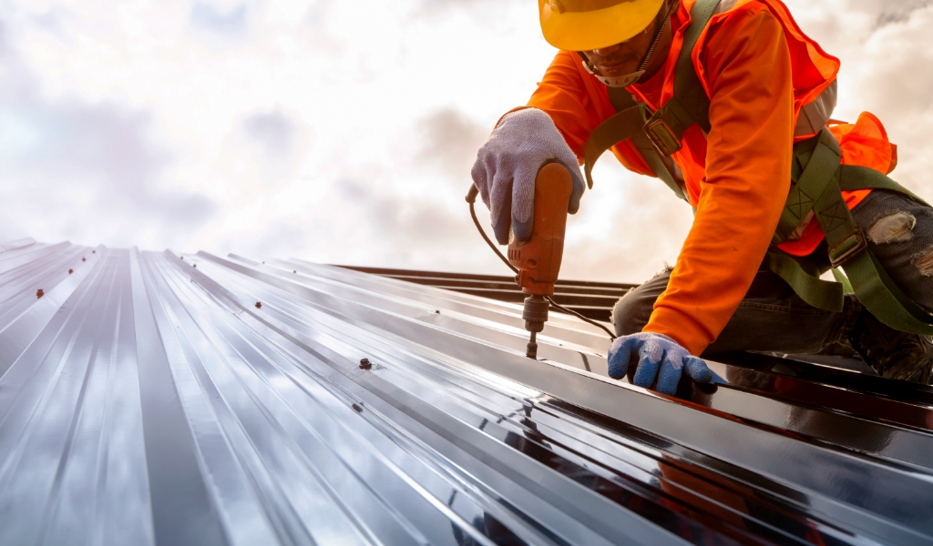 A worker in an orange safety vest and hard hat expertly uses a power drill on a metal roof under a cloudy sky, showcasing top-notch roofing services.