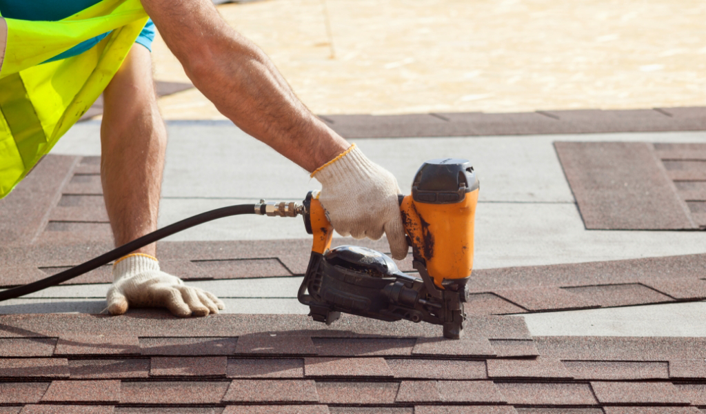 A worker from a roofing services team uses a nail gun to install asphalt shingles on a roof, ensuring safety with a reflective vest and gloves.