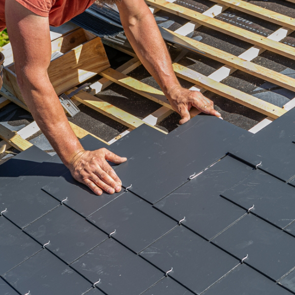 A person fitting dark slate tiles on a rooftop, surrounded by wooden beams and various roofing materials.