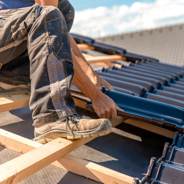 Under a clear sky, a person wearing work boots and jeans installs black roof tiles on a wooden frame, showcasing the expertise of professionals in the industry, much like you'd find with Marina Bay Roofing.