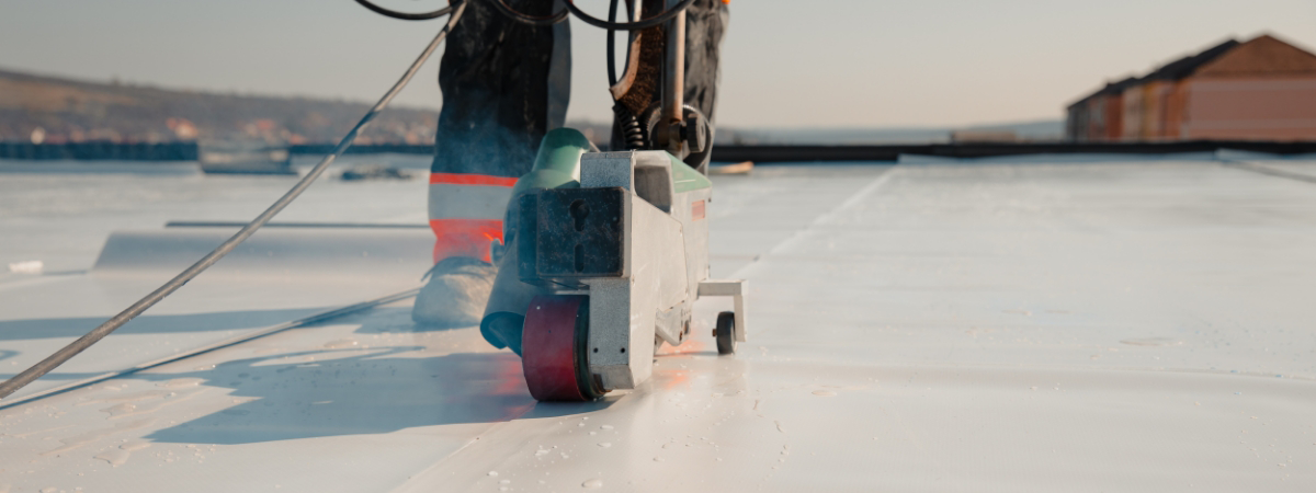 A person expertly operates a roofing machine on a flat surface, using quality roofing materials, with charming houses visible in the background.
