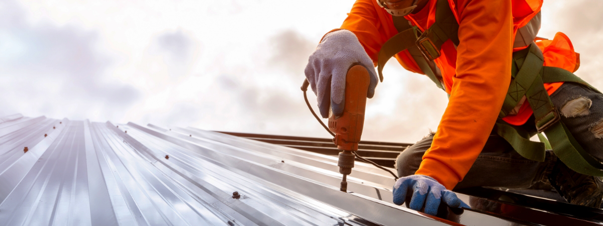 A construction worker in an orange safety vest and harness uses a drill on a metal roof with cutting-edge roofing materials, set against a cloudy sky.