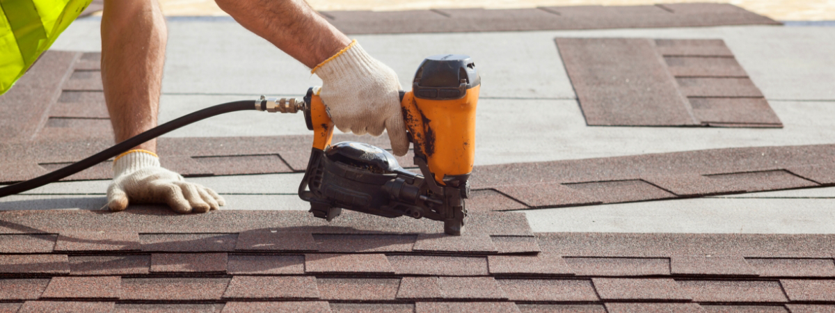 Person using a nail gun to install asphalt shingles, essential roofing materials, on a roof while wearing work gloves and a yellow safety vest.