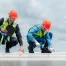 Two construction workers wearing orange helmets and safety vests inspect a roof; one uses a clipboard while the other examines the surface with a tool under a partly cloudy sky.