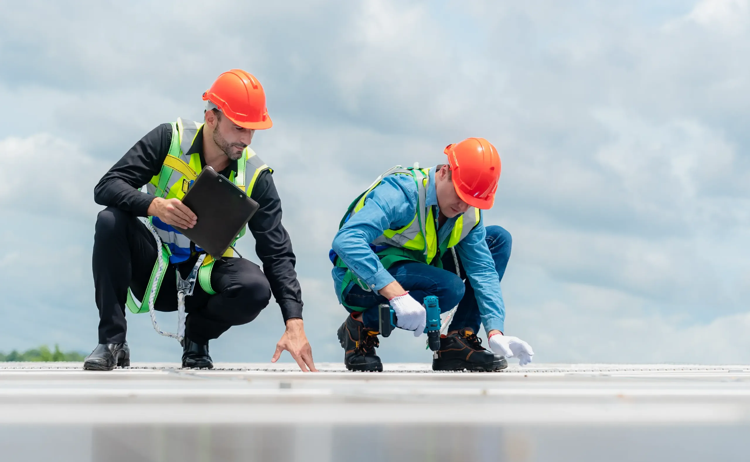 Two construction workers wearing orange helmets and safety vests inspect a roof; one uses a clipboard while the other examines the surface with a tool under a partly cloudy sky.