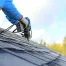 A roofer wearing safety gear uses a power drill to install asphalt shingles on a residential roof under a blue sky.